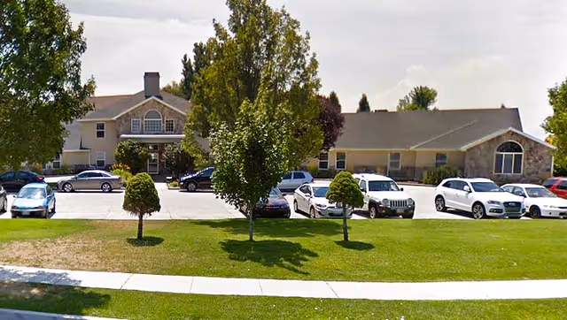Exterior view of a senior living facility named Bel Aire Senior Living showing a large building with multiple windows, a parking lot with several cars, green grass, and trees in the foreground under a partly cloudy sky.