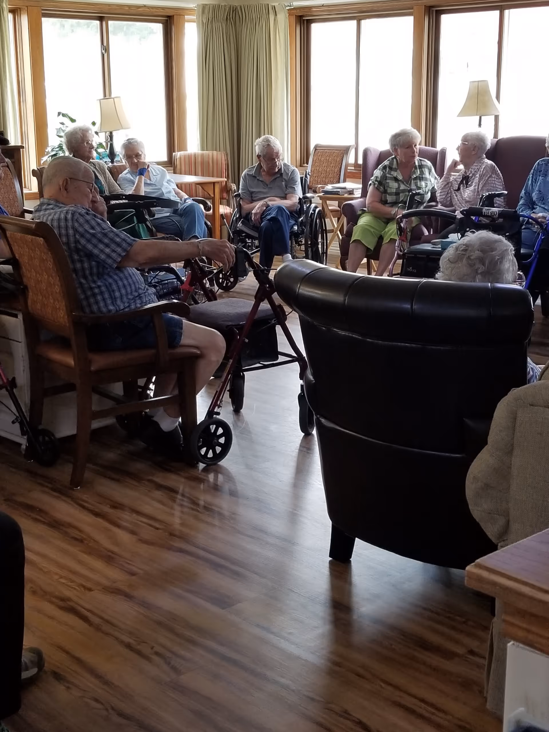 Several elderly residents sitting and chatting in a sunlit common living room with chairs, walkers, and large windows.