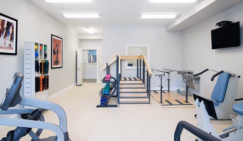 Bright interior therapy room with exercise machines, a set of practice stairs and parallel bars.