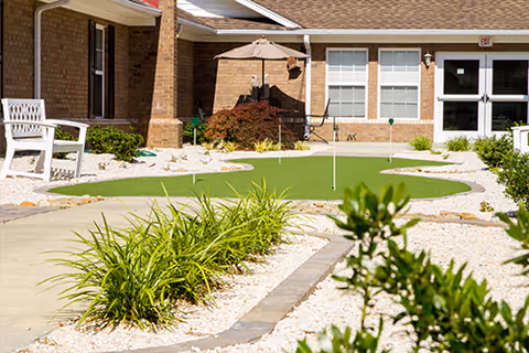 Outdoor courtyard area at a nursing and rehabilitation center featuring a small putting green, white gravel landscaping, green plants, a white bench, and brick building walls with windows and doors.