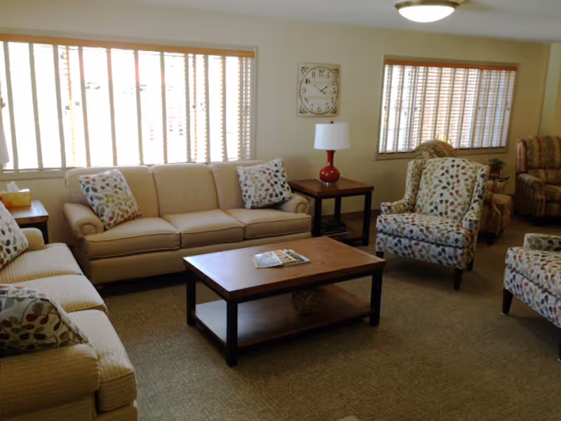 A well-lit living room with beige sofas and patterned armchairs arranged around a wooden coffee table. There are two large windows with blinds, a side table with a red lamp, and a wall clock above it.