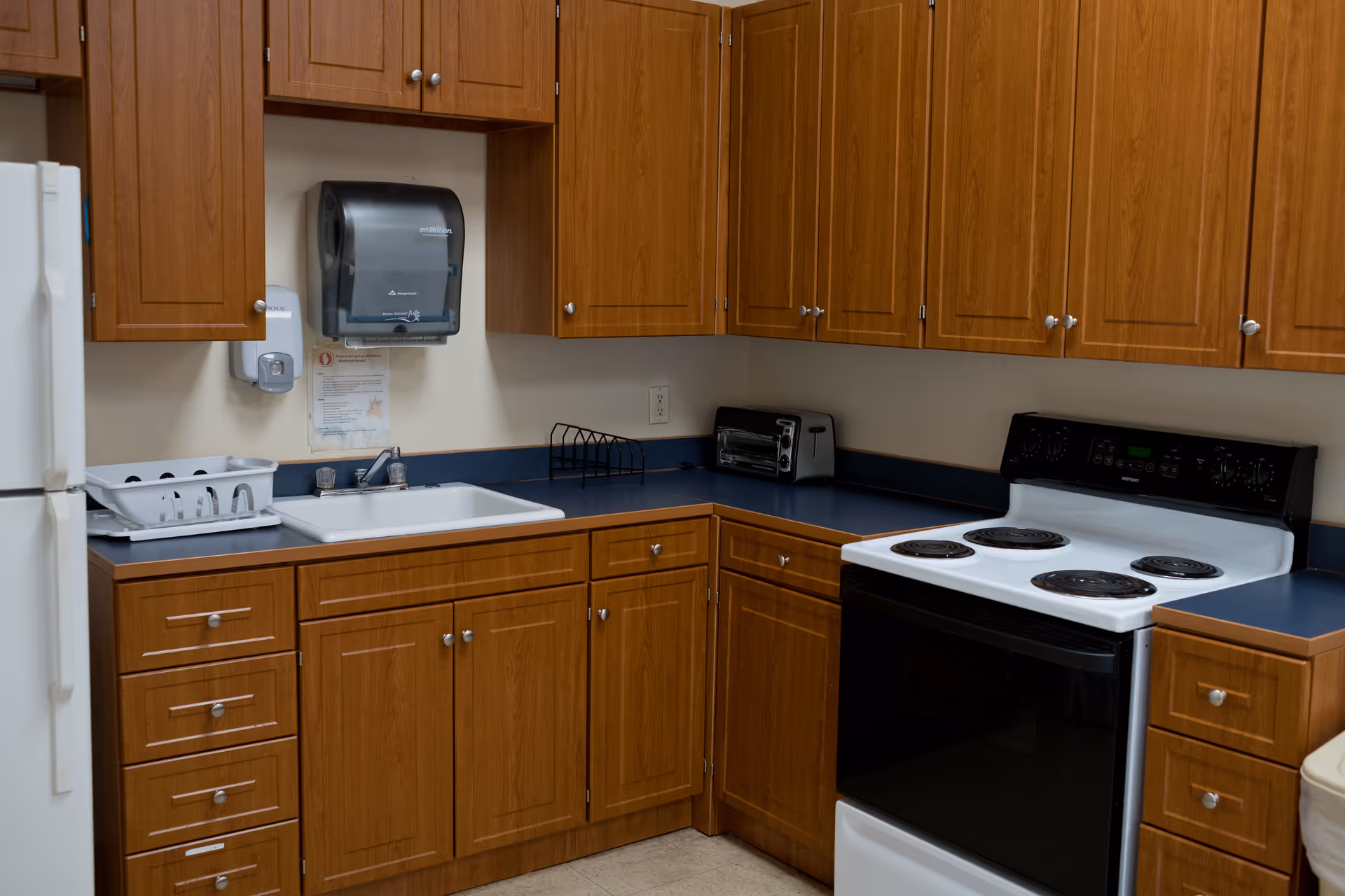 A kitchen area with wooden cabinets, a white refrigerator, a white electric stove with four burners, a sink with a dish rack, a toaster oven, and a paper towel dispenser mounted on the wall.