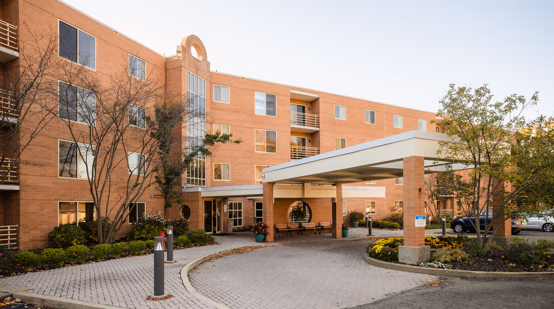 Exterior view of a multi-story brick building with large windows and a covered entrance driveway. There are trees and landscaped flower beds around the entrance area, and several parked cars are visible in the background.