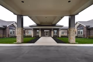 Covered entrance to a single-story brick building with stone pillars and a concrete walkway leading to glass doors. The building has multiple windows and a well-maintained lawn on either side of the walkway.