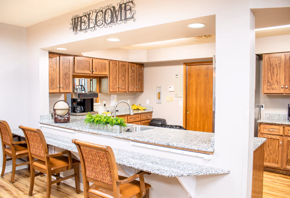 Bright communal kitchen with granite countertops, wooden cabinets, a sink, coffee maker and bar seating beneath a 'WELCOME' sign.