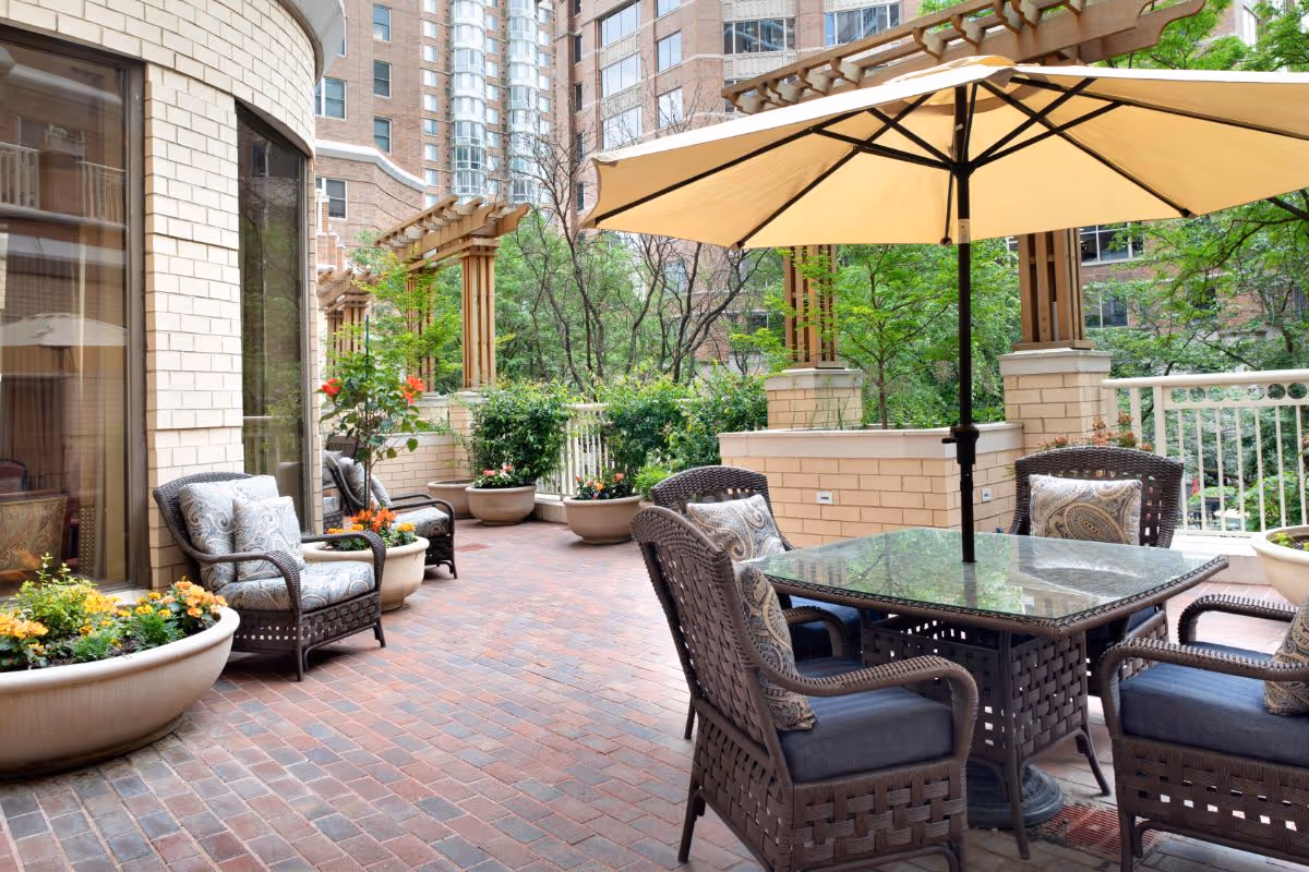 Outdoor patio at The Jefferson with wicker chairs around a glass-top table under a large umbrella, potted plants, and surrounding brick buildings.