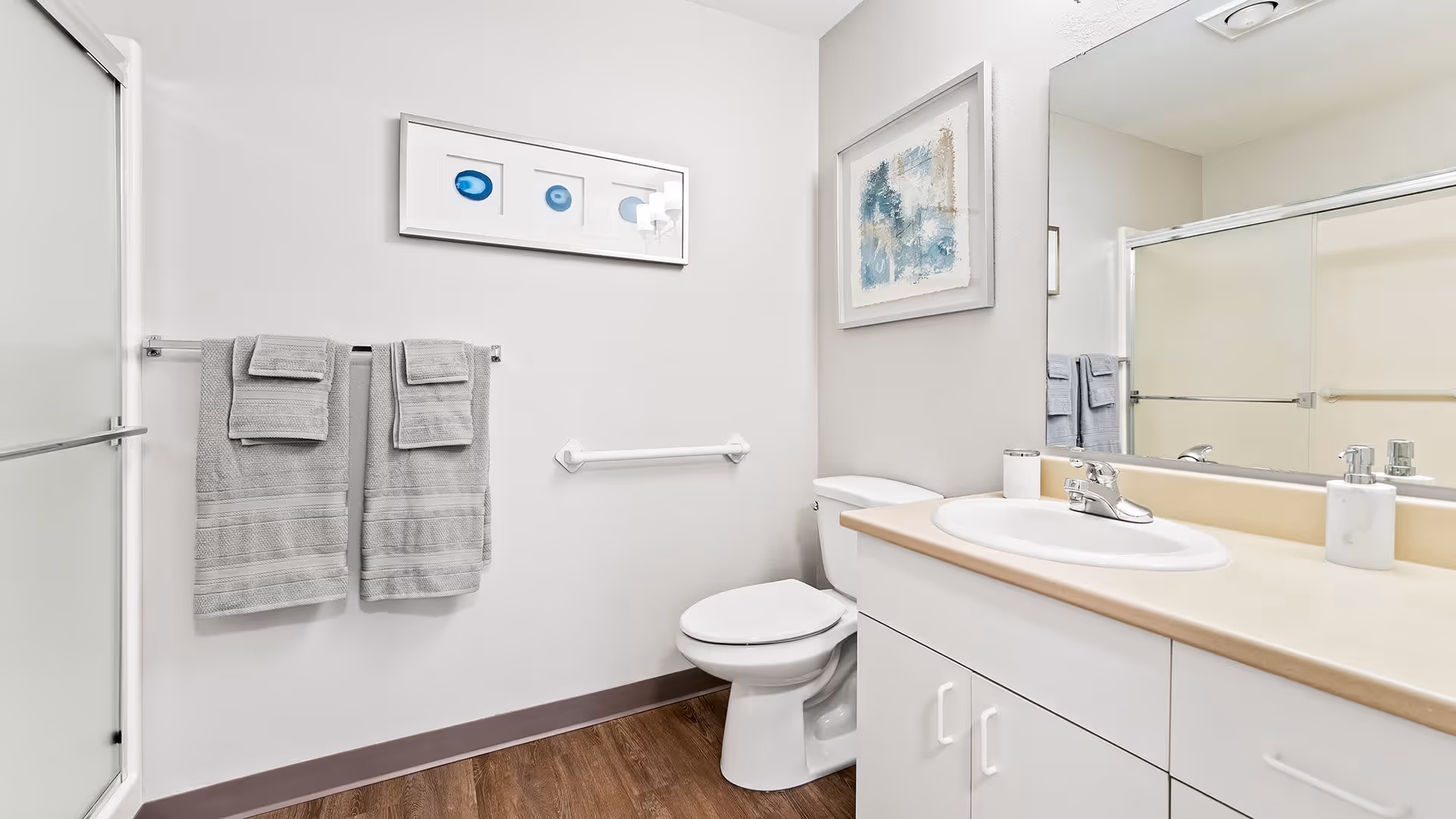 A clean and modern bathroom featuring a white toilet, a sink with a beige countertop, and a large mirror above the sink. Two gray towels hang on a towel rack next to a shower with sliding glass doors. The walls are white with two framed abstract artworks hanging above the toilet and towel rack. The floor has a wood-like finish.