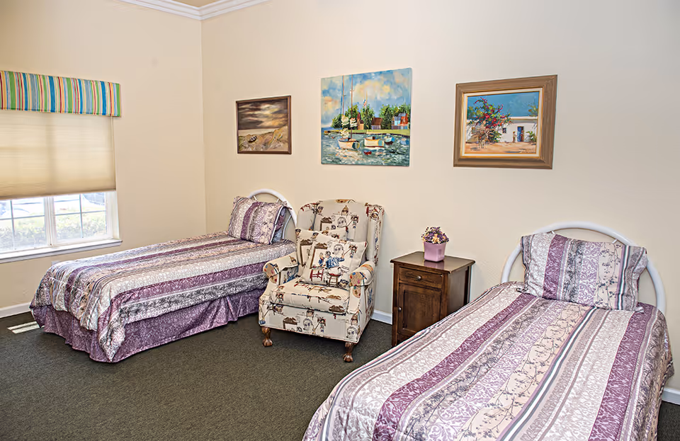 Two twin beds with purple patterned bedding, a patterned armchair and nightstand in a bright, simply furnished bedroom with wall art.