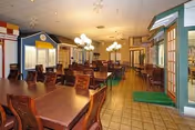 A spacious dining room with multiple wooden tables and chairs arranged neatly. The room features decorative streetlamp-style light fixtures and facades resembling small storefronts or houses along the walls, creating a quaint, village-like atmosphere.