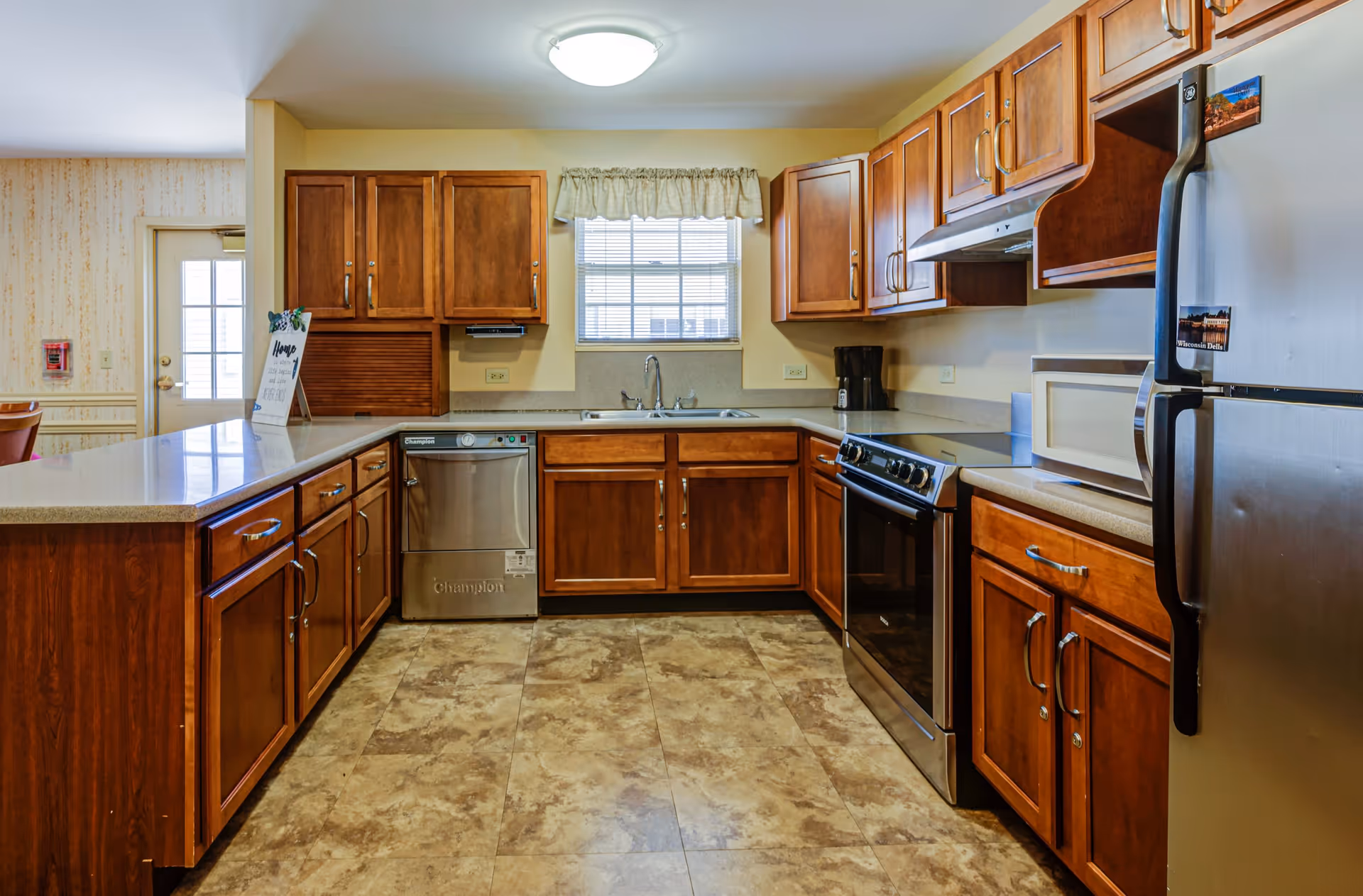 A clean, well-lit kitchen with wooden cabinets, a stainless steel dishwasher, stove, and refrigerator. There is a window above the sink with a small curtain, and a microwave is placed on the countertop. The floor has a tiled pattern, and there is a door leading to another room on the left side.