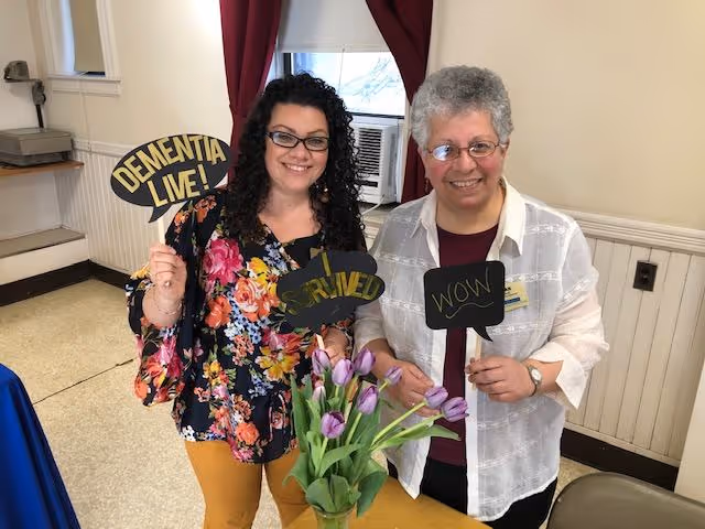 Two women standing indoors smiling and holding speech bubble signs that say 'DEMENTIA LIVE!', 'SURVIVED', and 'WOW'. There is a vase with purple tulips on a table in front of them, and a window with red curtains in the background.