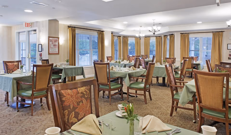 Dining room with multiple tables set with green tablecloths, wooden chairs, place settings, and large windows with gold curtains.