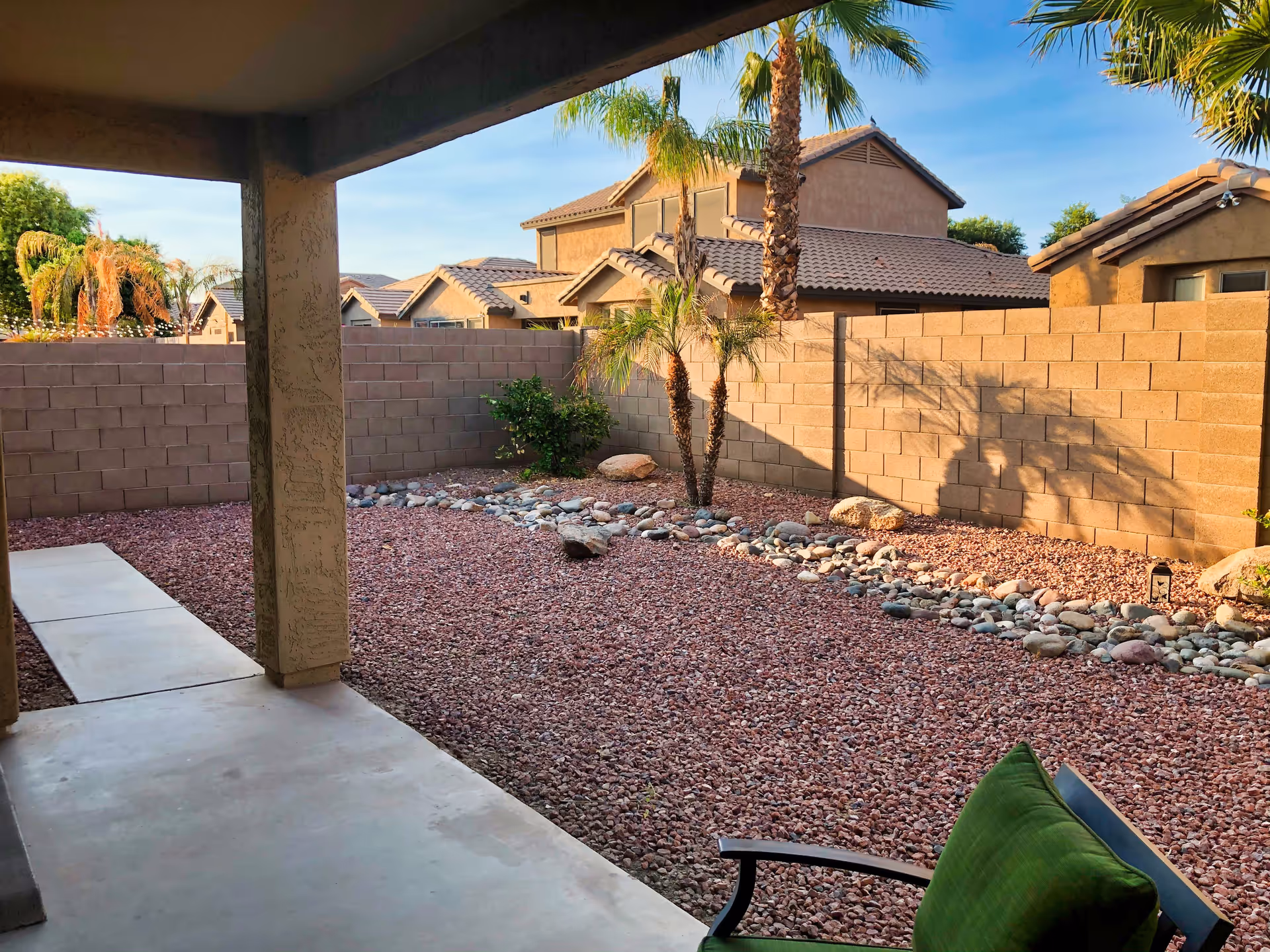 View of a backyard with a covered patio area, a concrete walkway, and a garden bed filled with red gravel and rocks. There are a few small palm trees and bushes along a beige brick wall, with neighboring houses visible in the background under a clear blue sky.