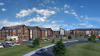Wide exterior view of a large, modern three-story senior living facility with multiple connected buildings, surrounded by green lawns, trees, and a parking lot with several cars under a partly cloudy sky.