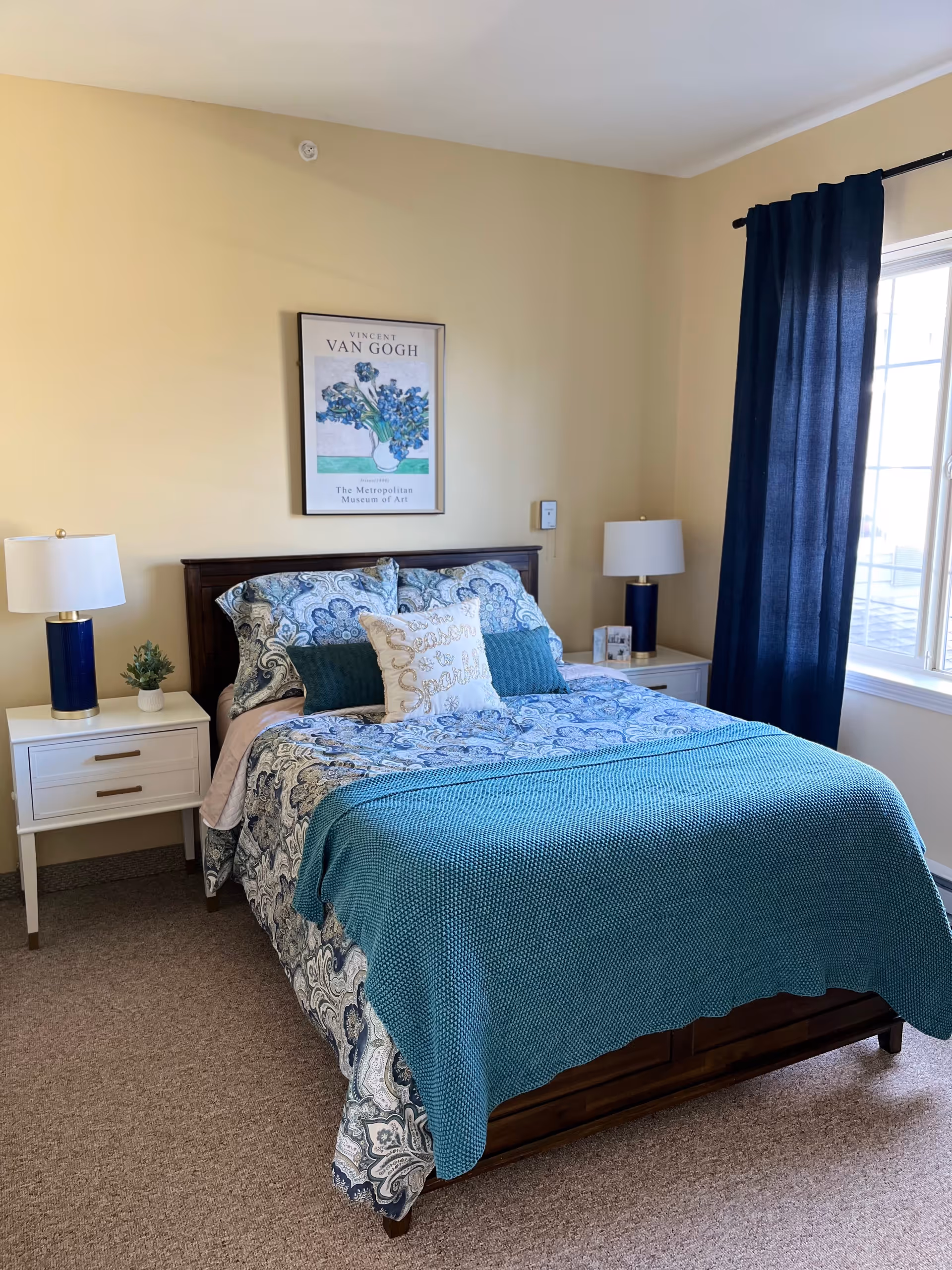 A neatly made bed with a blue and white patterned comforter and teal throw blanket in a bedroom. There are two white nightstands on either side of the bed, each with a blue lamp and one with a small plant. A framed Vincent Van Gogh art print hangs on the beige wall above the bed. A window with dark blue curtains is on the right side of the room.