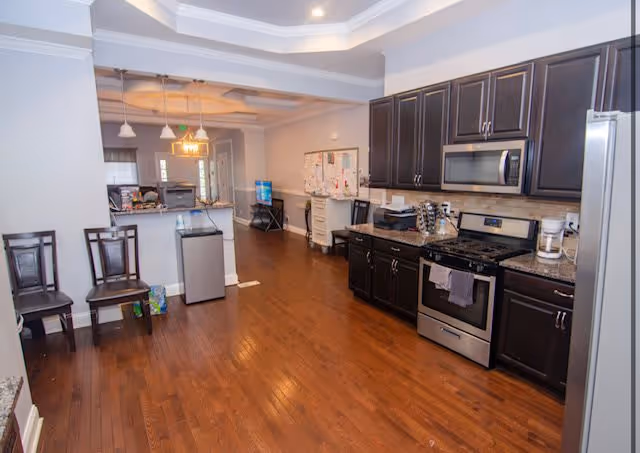 Interior view of a kitchen and dining area with dark wood cabinets, stainless steel appliances including a stove, microwave, and refrigerator. The floor is wooden, and there are two chairs against the wall on the left. Pendant lights hang over the counter area, and a bulletin board is visible on the far wall.