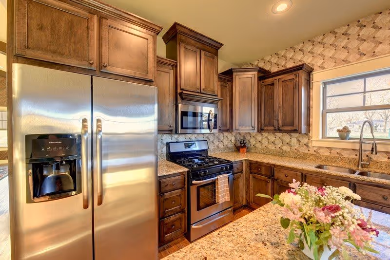A bright kitchen with wooden cabinets, a stainless steel refrigerator with a water and ice dispenser, a stainless steel gas stove with an oven, a microwave above the stove, granite countertops, a window above the sink, and a vase of flowers on the countertop.