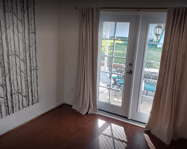 Sunlit interior room with hardwood floors, light curtains framing French doors to a patio, and tree-patterned wall art on the left.