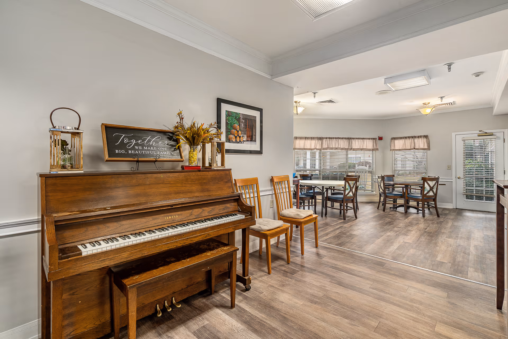 Interior view of a senior living facility common area featuring a wooden upright piano with decorative items on top, two wooden chairs with cushions, and a dining area with tables and chairs near windows with blinds and valances. The room has wood flooring and neutral-colored walls.