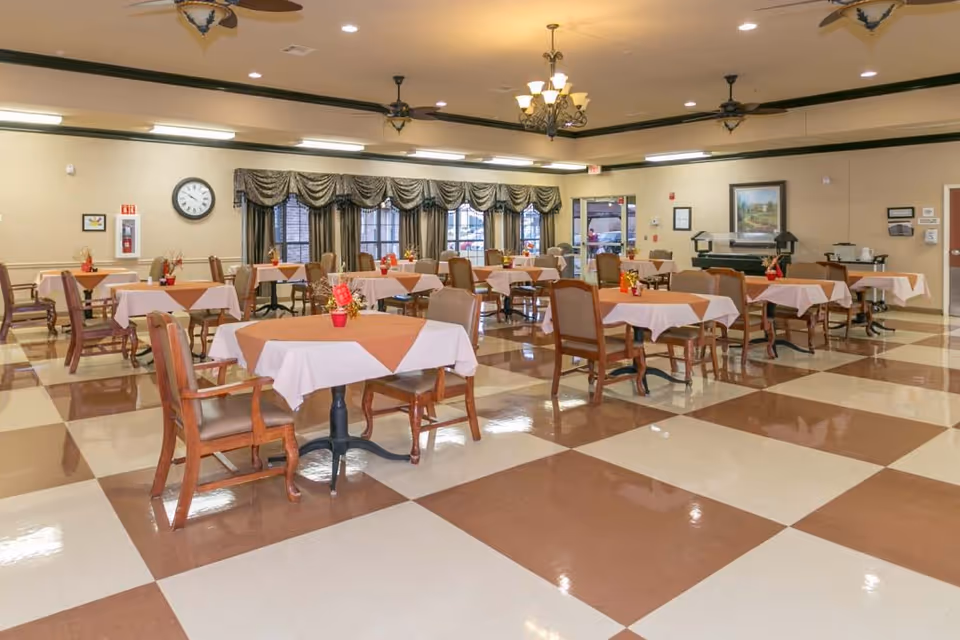 A spacious dining room with multiple tables covered in white and brown tablecloths, each table set with small red flower pots. The room has a checkered brown and cream tile floor, wooden chairs with armrests, large windows with draped curtains, ceiling fans, and chandeliers. There is a clock on the wall and a buffet area in the background.