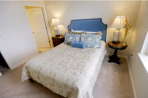 A neatly made bed with a blue upholstered headboard and decorative pillows in a bedroom. There are two bedside tables, each with a lamp. One bedside table is wooden with a drawer, and the other is round with a decorative vase. The room has beige walls and carpeted floor, with a door slightly ajar leading to another room.