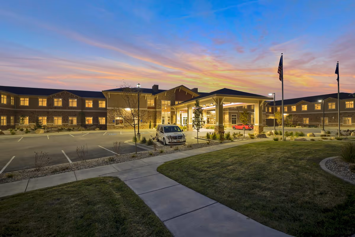 Dusk view of a senior living facility's front entrance with a covered porte-cochère, illuminated windows, parked cars, flags, and a landscaped walkway.