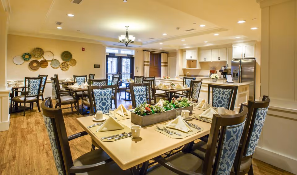 A well-lit dining room with multiple tables set with napkins, cups, and silverware. The chairs have blue patterned upholstery. The room features wooden flooring, a decorative wall with hanging plates, and a kitchen area with white cabinets and stainless steel appliances in the background.