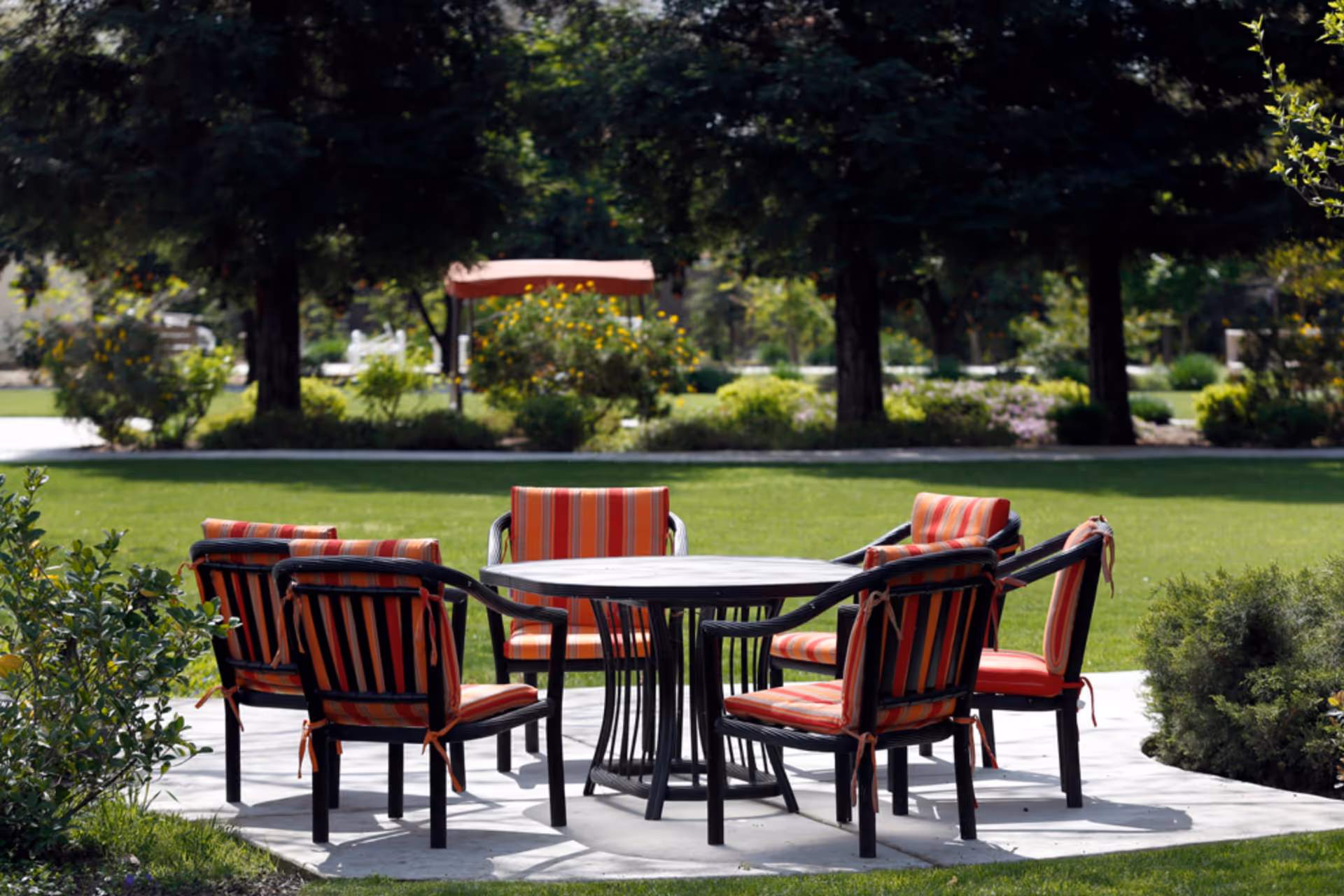 Round outdoor table surrounded by six striped cushioned chairs on a patio in a grassy garden with trees in the background.