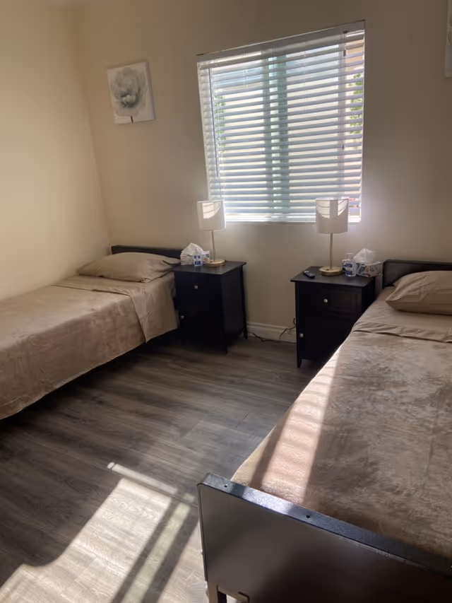 A simple bedroom with two single beds placed parallel to each other, each with beige bedding and a pillow. Between the beds are two black nightstands, each with a lamp, a box of tissues, and a cup. A window with white blinds lets in natural light, and a small floral painting hangs on the wall above one bed. The floor is covered with wood-patterned vinyl or laminate.