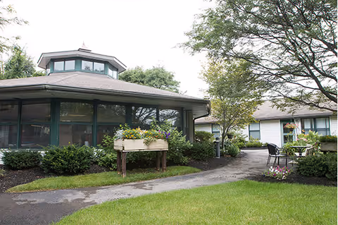 Outdoor view of a senior living facility with a paved walkway, green grass, bushes, and trees. There is a round building with large windows and a smaller building in the background. Outdoor seating with chairs and a table is visible near the walkway.