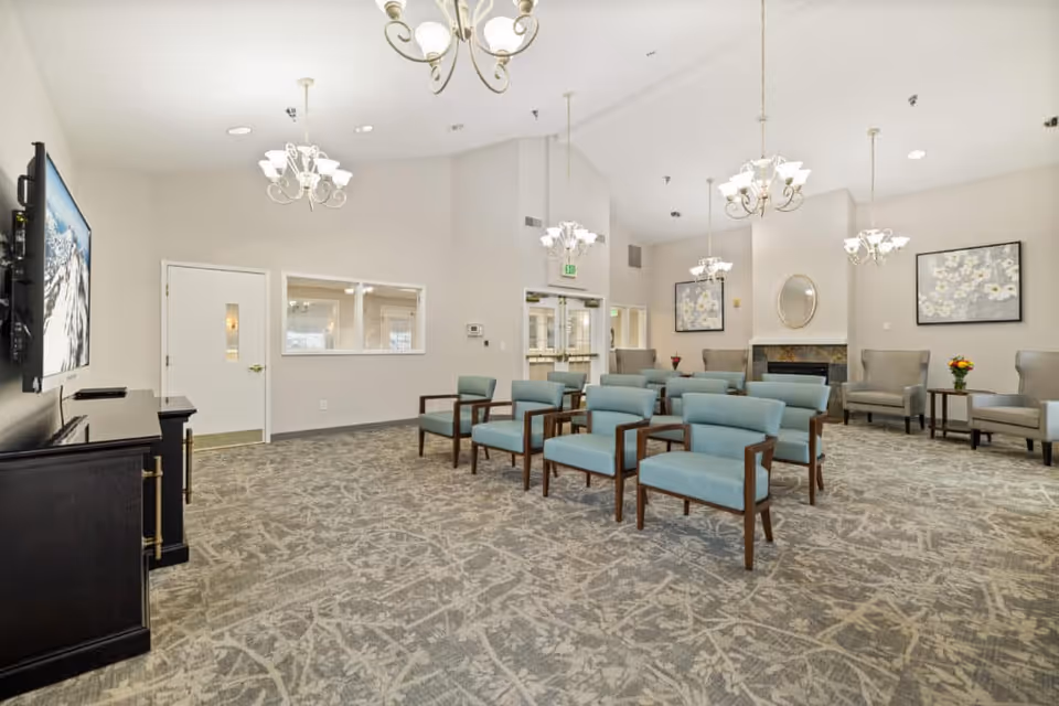 Community seating room with rows of blue upholstered chairs, chandeliers, a TV on the left, and a fireplace on the far wall.