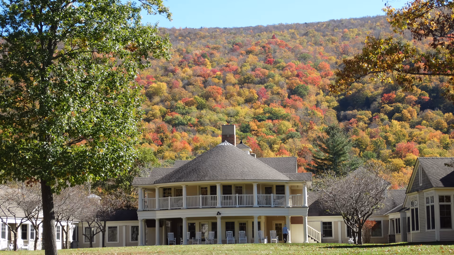 A large two-story building with a wraparound porch and multiple white rocking chairs on the lower level porch. The building is surrounded by trees, some with autumn foliage, and is set against a backdrop of a forested hillside with colorful fall leaves in shades of red, orange, and yellow under a clear blue sky.