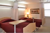 A shared nursing facility bedroom with two hospital-style beds, maroon bedspreads, overbed tables, a bedside lamp, and curtain dividers.
