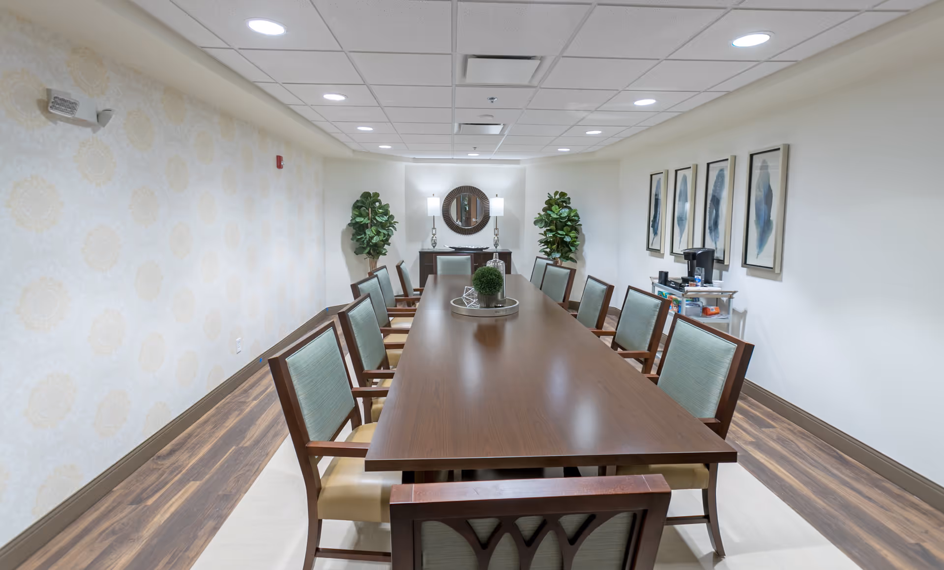 A well-lit conference or dining room with a long wooden table surrounded by ten chairs with light green upholstery. The room has a patterned wallpaper on one side and plain white walls on the other, decorated with four framed abstract art pieces. At the far end, there is a sideboard with a round mirror above it, flanked by two lamps and two potted plants. A small cart with a coffee maker and supplies is visible on the right side.