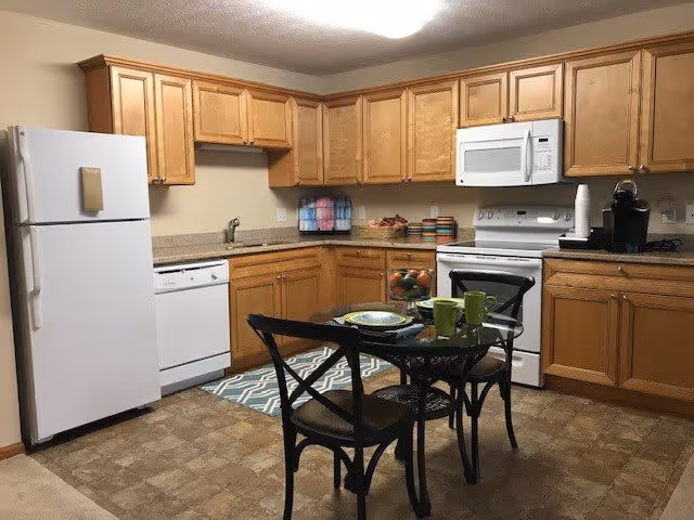 A kitchen with wooden cabinets, a white refrigerator, dishwasher, stove, and microwave. There is a small glass dining table with two chairs, set with plates and green mugs. The floor has a patterned linoleum surface and a small rug is placed near the sink.