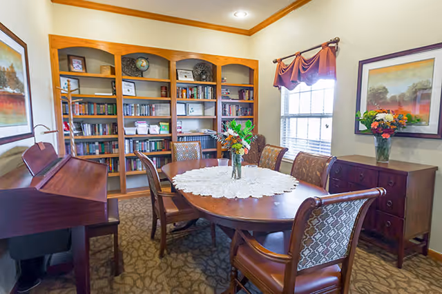 A cozy room with a wooden oval table surrounded by six upholstered chairs. A vase with flowers sits on a lace tablecloth in the center of the table. Behind the table is a large wooden bookshelf filled with books and decorative items. To the left, there is a wooden piano with a bench. On the right side, a window with a valance lets in natural light, and below it is a wooden sideboard with a vase of flowers. The walls are decorated with framed artwork.