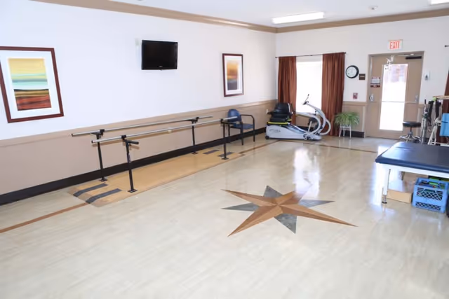 Interior rehabilitation room with parallel bars, an exercise bike, treatment table, wall-mounted TV and a decorative star inlaid on the floor.