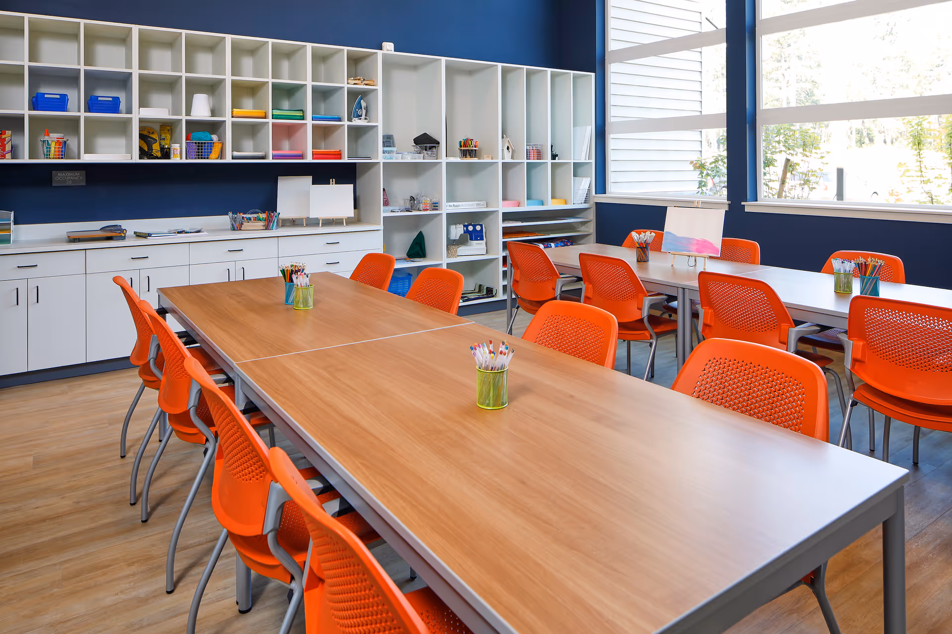 A bright and organized craft room with two long wooden tables surrounded by orange chairs. The room features white cubby shelves filled with various craft supplies, including colored paper, markers, and an iron. Large windows allow natural light to fill the space.