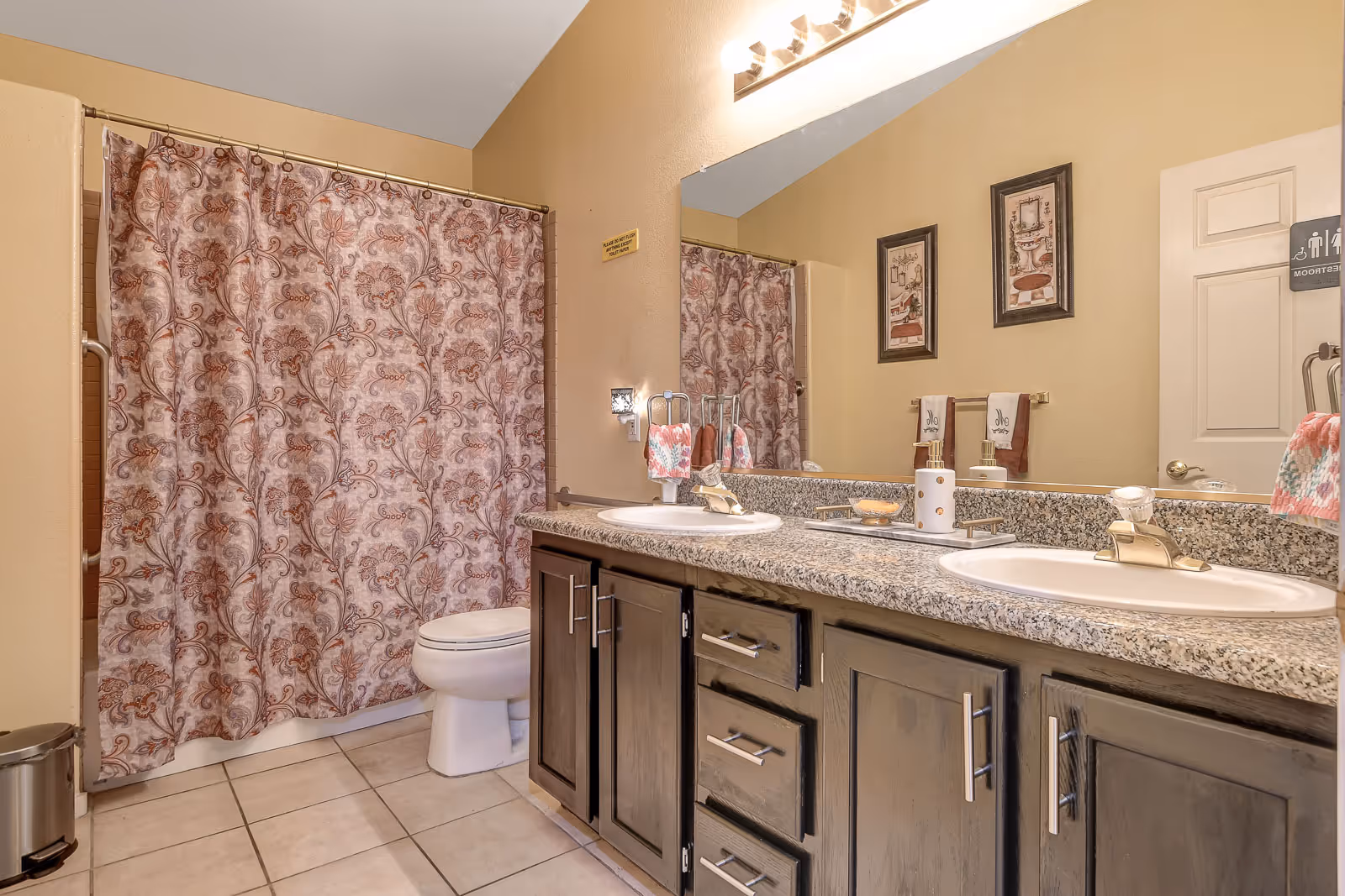 A bathroom with a double sink vanity featuring granite countertops and dark wood cabinets. There is a toilet next to a bathtub with a floral patterned shower curtain. The walls are painted beige, and there are two framed pictures above the towel rack. A large mirror spans the length of the vanity, reflecting the bathroom door with a restroom sign.