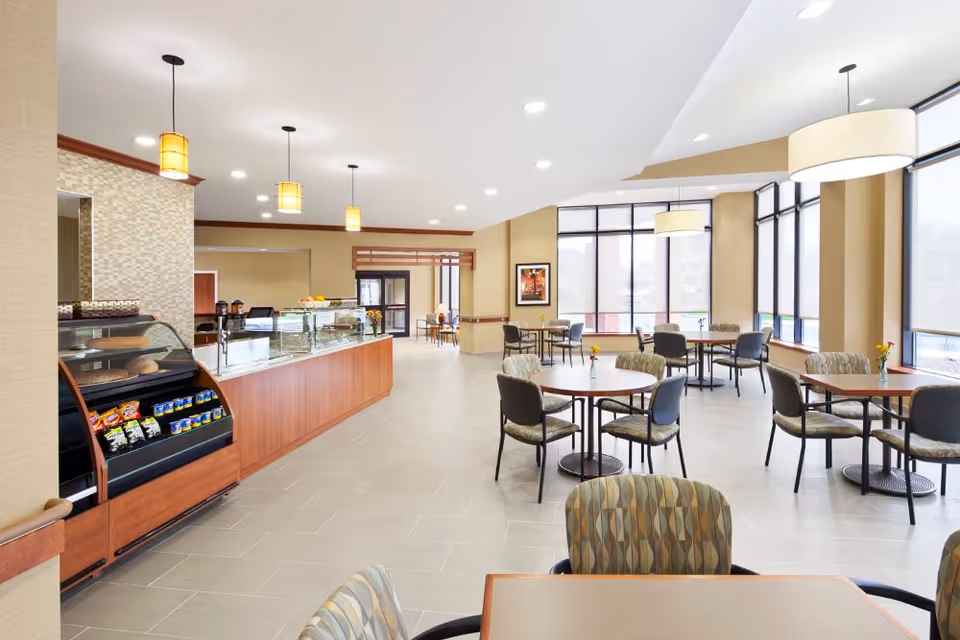 Bright cafeteria-style dining area with round tables and chairs, large windows, and a service counter/display case.