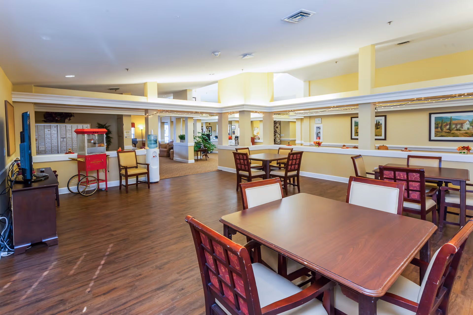 Communal dining room with several wooden tables and chairs, a popcorn cart, TV and seating area in a senior living facility.
