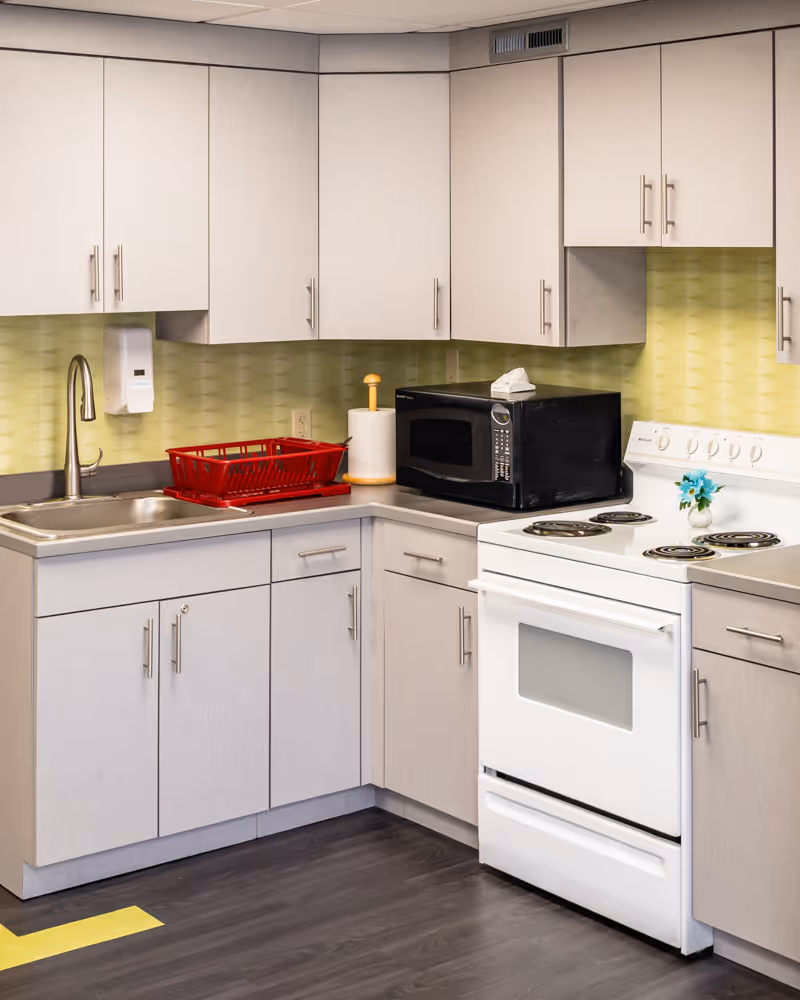 A clean kitchen corner with light gray cabinets, a stainless steel sink with a faucet, a red dish drying rack, a paper towel holder, a black microwave, and a white electric stove with four coil burners. The backsplash is light green with a textured pattern, and there is a small vase with blue flowers on the stove. The floor is dark wood, and there is a yellow tape marking on the floor.
