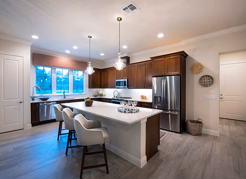Bright modern kitchen with a large white island, three upholstered barstools, dark wood cabinets, and stainless steel appliances.