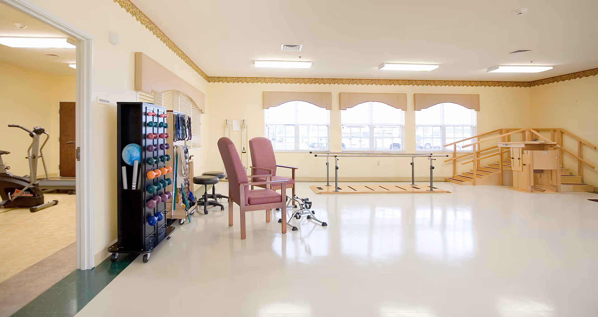 A spacious physical therapy or rehabilitation room with exercise equipment including parallel bars, a ramp with handrails, chairs, and a rack of colorful dumbbells. Adjacent room visible with additional exercise machines.
