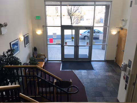 Lobby of a senior living facility with a stair railing, seating area, indoor plants, and glass double doors leading outside.
