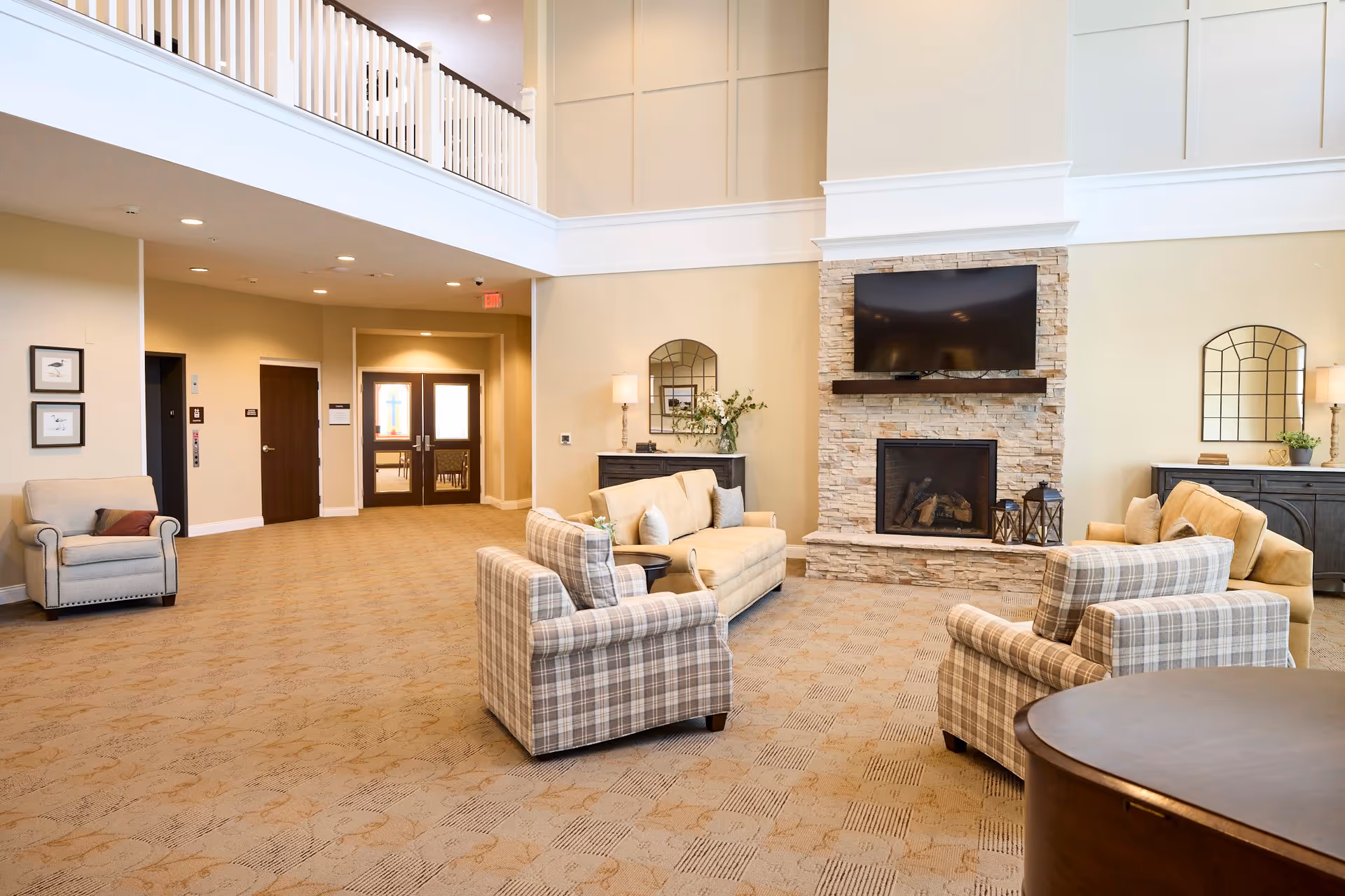 A spacious and well-lit living room area with beige walls and carpeted floor. The room features a stone fireplace with a mounted flat-screen TV above it, two plaid armchairs, and two beige sofas arranged around a small round table. There are decorative mirrors and lamps on dark wooden sideboards against the walls, and a balcony railing is visible on the upper level.