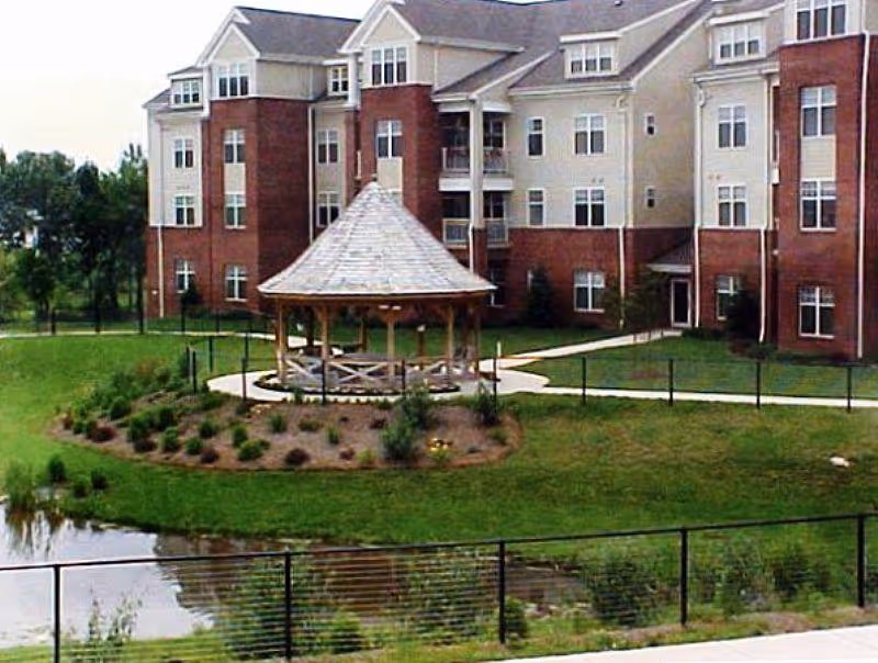 Exterior view of a multi-story senior living building with a gazebo by a pond and landscaped grounds.