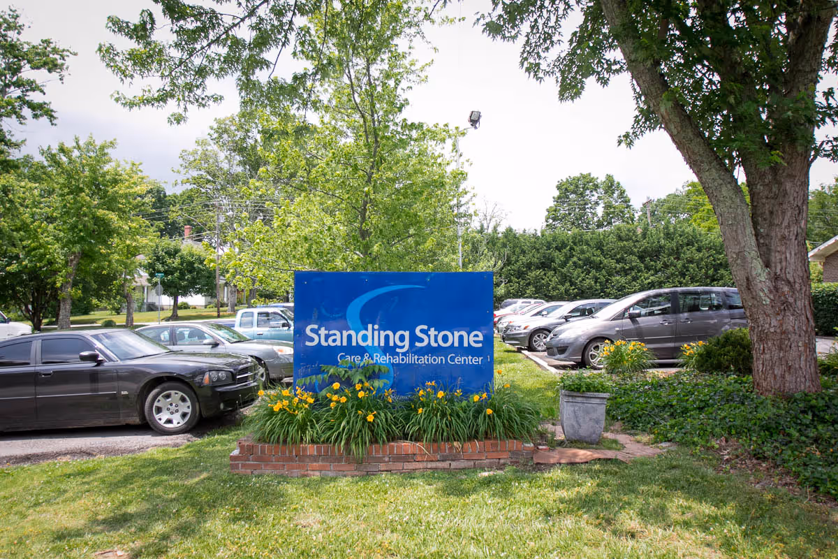 Outdoor view of the parking area at Standing Stone Care & Rehabilitation Center with a blue sign displaying the facility's name surrounded by green grass, yellow flowers, trees, and several parked cars.