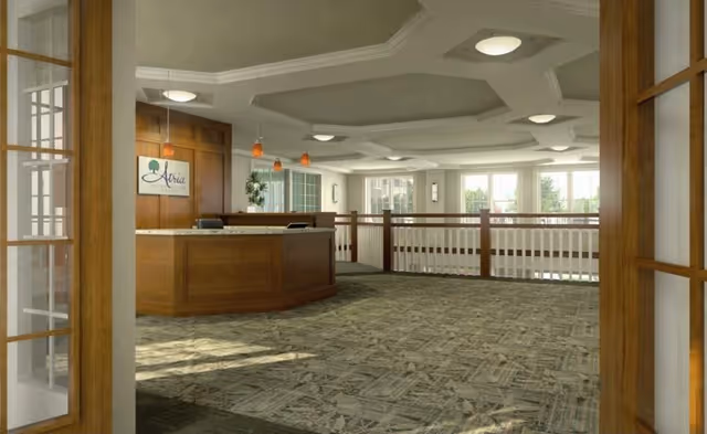Interior view of a senior living facility reception area with a wooden front desk, pendant lights hanging above, large windows letting in natural light, and a patterned carpet floor. The ceiling has decorative molding and recessed lighting, and there are wooden framed glass doors partially open in the foreground.