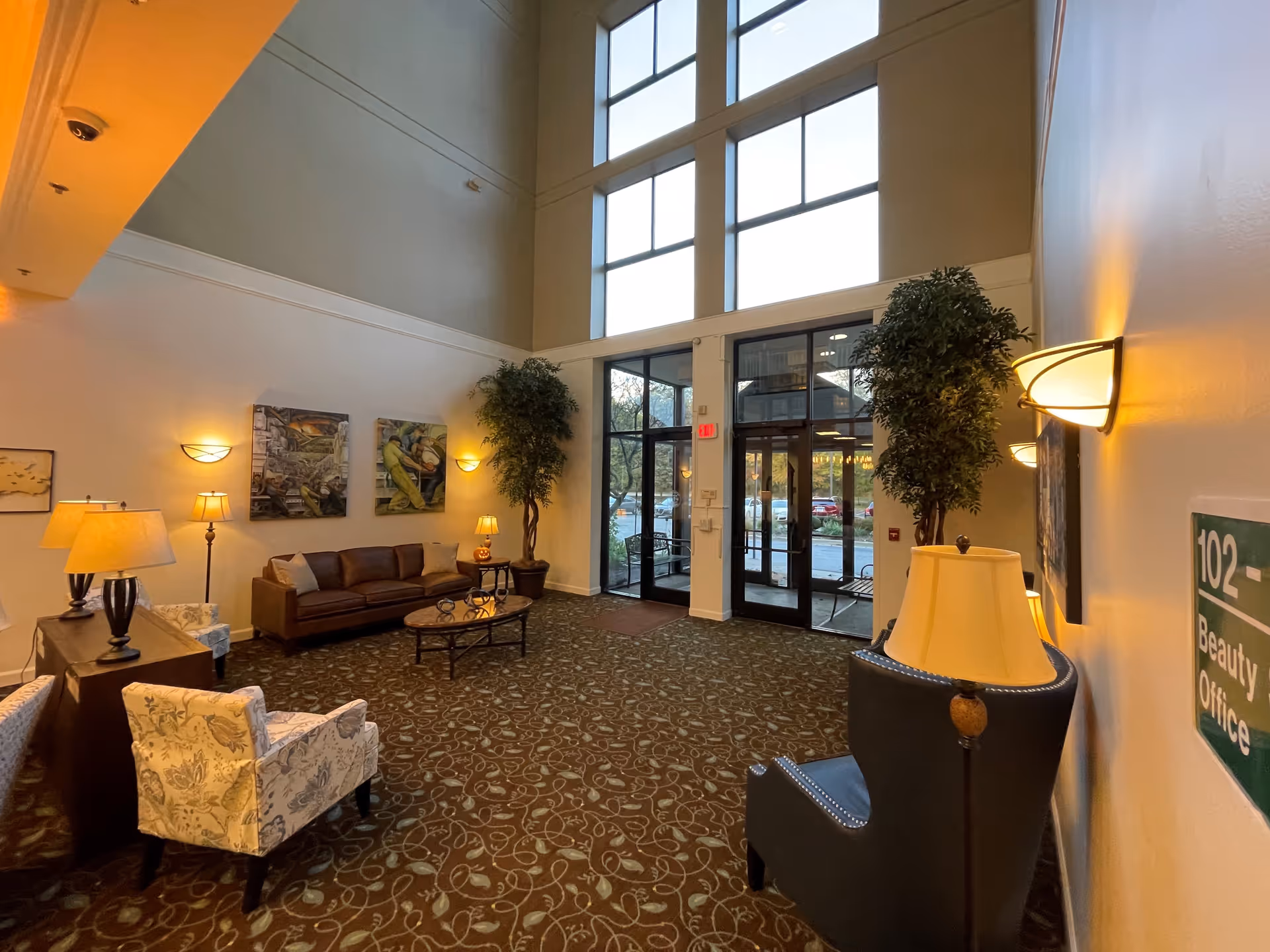 A spacious and well-lit lobby area with high ceilings and large windows. The room features a brown leather sofa, patterned armchairs, a coffee table, several lamps, potted plants, and wall art. There is a glass entrance door with a view of parked cars outside. A sign on the wall indicates '102 Beauty Office'.